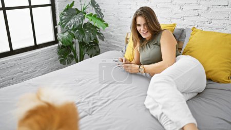 Téléchargez les photos : Souriante jeune femme hispanique couchée au lit avec son chien bien-aimé, utilisant joyeusement son smartphone dans le confort douillet de sa chambre - en image libre de droit