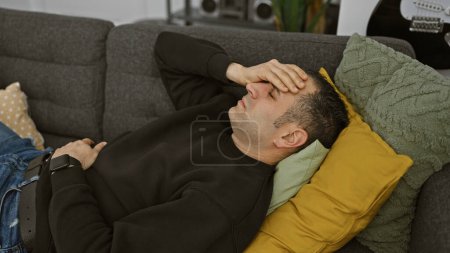 Ein gestresster junger hispanischer Mann liegt drinnen auf einer Couch, die Hand auf der Stirn und drückt Erschöpfung oder Kopfschmerzen aus.