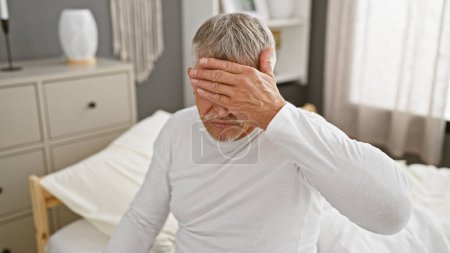 Photo for A distressed senior man with grey hair sitting in a bedroom, covering his face with his hand. - Royalty Free Image