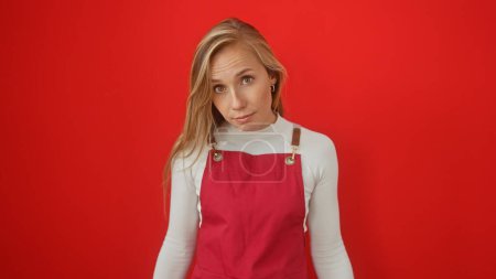 Blonde woman in white top and red apron posing against an isolated red background looks thoughtfully at the camera