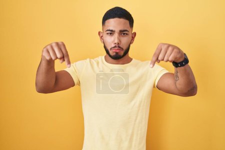 Young hispanic man standing over yellow background pointing down looking sad and upset, indicating direction with fingers, unhappy and depressed. 