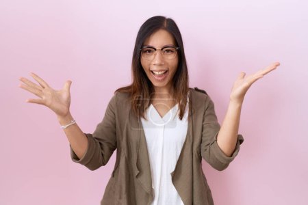 Foto de Mujer china de mediana edad con gafas sobre fondo rosa celebrando loco y sorprendido por el éxito con los brazos levantados y los ojos abiertos gritando emocionado. concepto ganador - Imagen libre de derechos