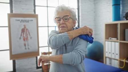 Eine Seniorin klebt in einer Physiotherapie-Klinik Eis auf ihre Schulter mit anatomischen Plakaten, die für Gesundheit stehen.