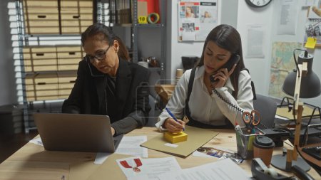 Two women investigators work diligently in a cluttered office, one on the phone taking notes, the other focused on her laptop.