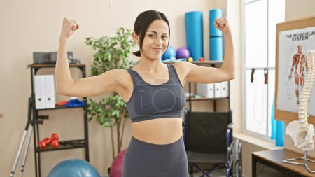 A confident hispanic woman flexes her muscles in a well-equipped rehabilitation clinic.