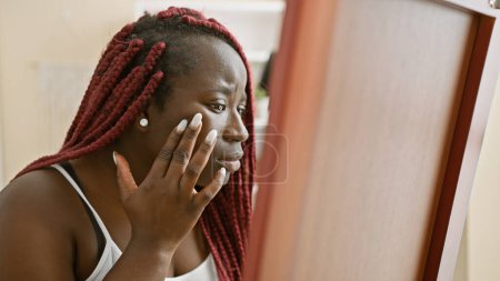 Photo for A contemplative african american woman with braids gazes at her reflection in a mirror at home. - Royalty Free Image
