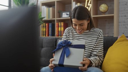 Photo for A cheerful hispanic woman enjoys unwrapping a gift in a cozy, book-filled living room. - Royalty Free Image