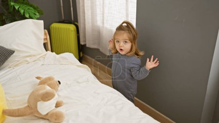 Téléchargez les photos : Surprise fille blonde debout dans une chambre avec un jouet en peluche et valise indiquant Voyage ou vacances. - en image libre de droit