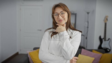 Téléchargez les photos : Souriant jeune femme caucasienne avec des lunettes posant judicieusement dans un salon confortable avec une guitare en arrière-plan - en image libre de droit