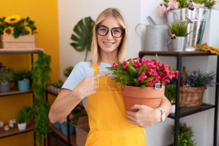 Foto de Young caucasian woman working at florist shop holding plant smiling happy pointing with hand and finger - Imagen libre de derechos
