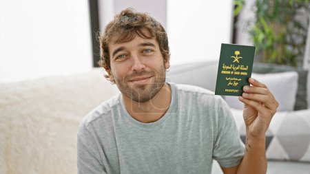 Confident, happy young arab man, lounging on the sofa at home, grinning ear to ear, while clutching his saudi arabian passport, ready to enjoy his next foreign holiday!