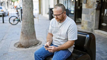 Attraktiver kaukasischer Mann mittleren Alters mit ernstem Gesichtsausdruck, der eifrig eine Nachricht auf sein Smartphone tippt, während er draußen auf einer Stadtbank im sonnigen Sonnenlicht sitzt.