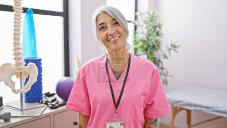 Foto de La sonriente y confiada fisioterapeuta de cabello gris de mediana edad disfruta alegremente de su trabajo, de pie en el corazón de una bulliciosa clínica de rehabilitación. - Imagen libre de derechos