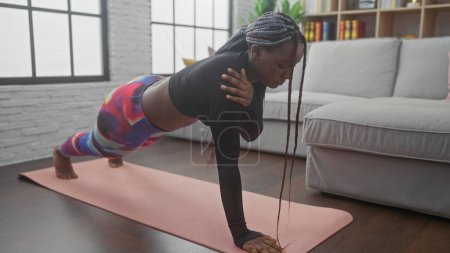 Téléchargez les photos : Femme afro-américaine avec des tresses effectue push-up sur tapis de yoga dans son appartement salon - en image libre de droit