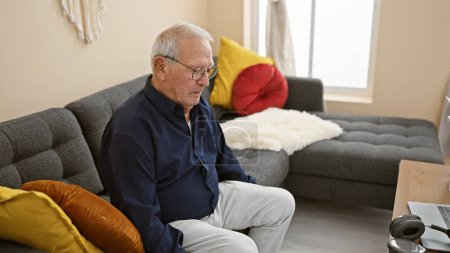 Serious faced senior man with white hair, comfortably sitting on a cozy sofa, relaxing at home with his eyes closed, a portrayal of mature relaxation in the comfort of indoor living room.