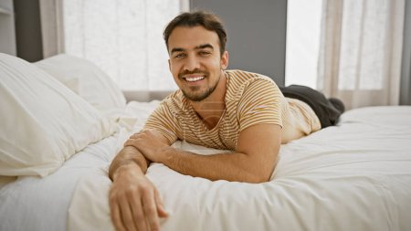 Foto de Hombre hispano guapo sonriendo mientras se relaja solo en un interior de dormitorio moderno bien iluminado - Imagen libre de derechos