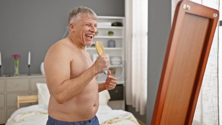 Joyful shirtless senior man singing with a hairbrush in front of a mirror in a cozy bedroom setup.