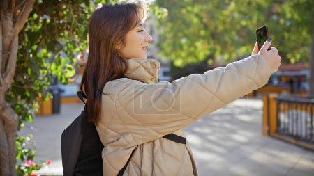 Photo for Young woman capturing a selfie in a sunny park using her smartphone, exuding casual elegance and joy. - Royalty Free Image