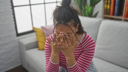 Photo for A young, distressed hispanic woman covering her face with hands, sitting on a white sofa in a bright, modern indoor setting. - Royalty Free Image