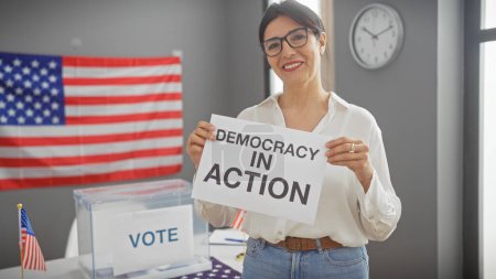 Smiling woman holding sign saying 'democracy in action' with us flag and ballot box indicating voting in america.