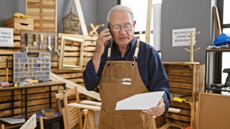Photo for Mature man hunched over documents in his timber-laden carpentry workshop, chin perched atop glasses as he chats away on his smartphone, mid-conversation amidst his lifelong woodwork profession. - Royalty Free Image
