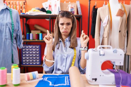 Mujer joven hispana modista diseñadora en la sala del taller apuntando hacia arriba buscando triste y molesto, indicando la dirección con los dedos, infeliz y deprimido. 
