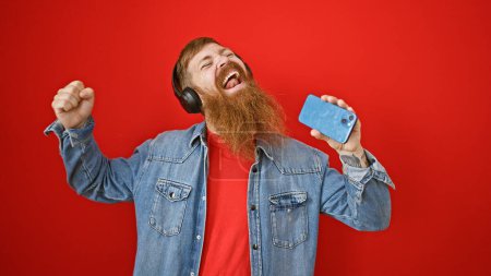 Foto de Joven pelirrojo confiado, sonriendo con auriculares, cantando una canción escuchando música sobre su teléfono móvil, de pie sobre un fondo rojo aislado - Imagen libre de derechos