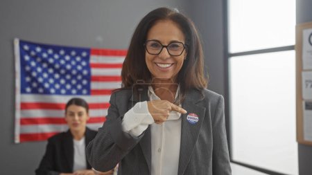 Photo for Smiling woman with 'i voted' sticker pointing at herself in front of another woman and american flag, indoors - Royalty Free Image