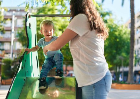 Téléchargez les photos : Mère et fils souriant confiants jouer sur la glissière au parc aire de jeux - en image libre de droit
