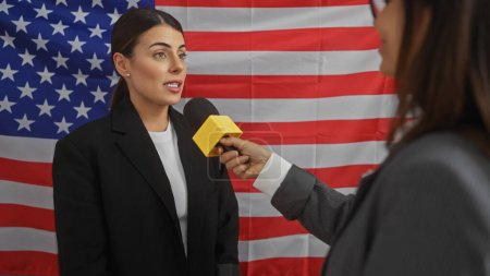 Professional woman interviewed by journalist in room with american flag backdrop.