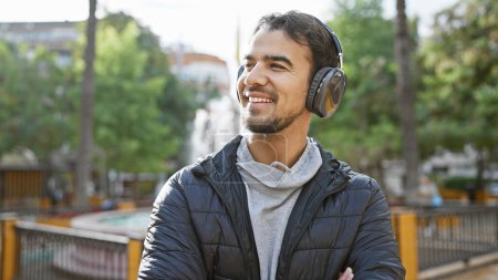 Foto de Hombre hispano sonriente con auriculares disfrutando de la música en un frondoso parque de la ciudad. - Imagen libre de derechos