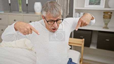 Photo for Surprised silver-haired man in glasses pointing at himself indoors, with bedroom furniture in the background. - Royalty Free Image