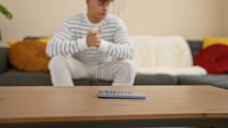 Besorgter junger hispanischer Mann wartet nervös auf ein Smartphone, zu Hause auf dem Sofa sitzend, ein Porträt der Angst in seinem hübschen Gesichtsausdruck.