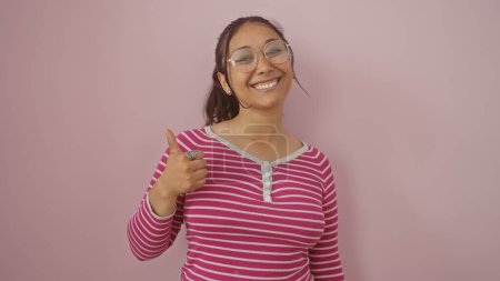 Téléchargez les photos : Jeune femme hispanique souriante donnant les pouces vers le haut, isolée sur un fond de mur rose, dépeignant la positivité et la confiance. - en image libre de droit