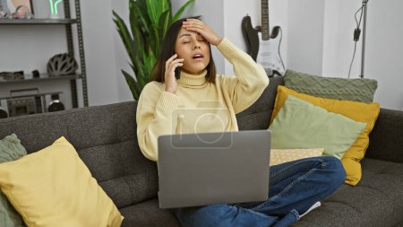 Téléchargez les photos : Femme hispanique stressée avec ordinateur portable et téléphone sur canapé dans un salon moderne - en image libre de droit