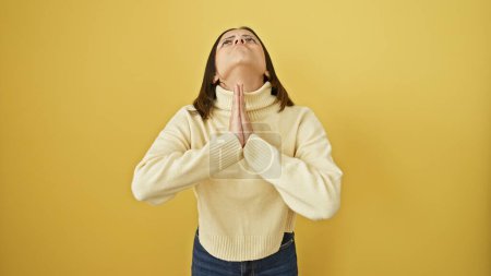 A contemplative young hispanic woman looks upwards, hands clasped in prayer against a vivid yellow background.
