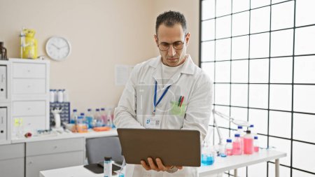 Un hombre hispano enfocado en un entorno de laboratorio examina los datos de su computadora portátil en medio de equipos médicos y cristalería.