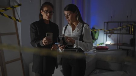 Photo for Two women investigators examine evidence using smartphones in the dimly lit room of a house crime scene. - Royalty Free Image
