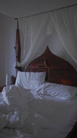 Téléchargez les photos : Une chambre faiblement éclairée avec un lit en bois complexe et des rideaux transparents créant une ambiance calme et sereine. - en image libre de droit