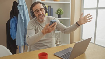 Ein fröhlicher Mann mittleren Alters mit Bart telefoniert zu Hause mit einem Smartphone, Kopfhörer und Laptop.