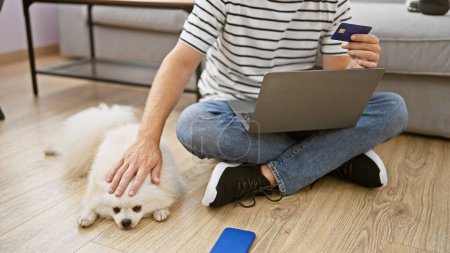 Téléchargez les photos : Jeune homme caucasien avec chien utilisant ordinateur portable et carte de crédit assis sur le canapé à la maison - en image libre de droit
