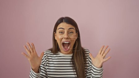 Photo for A cheerful young hispanic woman in a striped shirt expressing excitement against a pink isolated background. - Royalty Free Image
