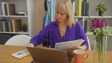 Foto de Una mujer rubia enfocada lee documentos en una oficina en casa con una computadora portátil, flores y taza de café sobre la mesa. - Imagen libre de derechos