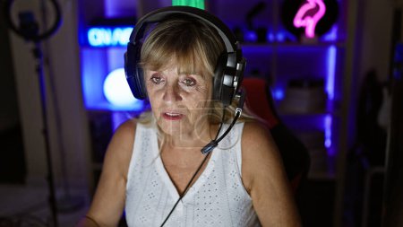 Photo for Serious-faced, middle-aged blonde woman streamer, rocking headphones in a dark gaming room, engrossed in immersive virtual entertainment - Royalty Free Image