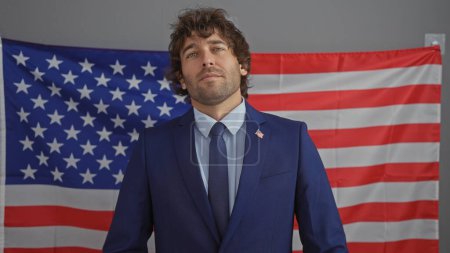 Foto de Hombre hispano guapo en traje con barba posando frente a la bandera americana en el interior, exudando confianza y profesionalismo. - Imagen libre de derechos
