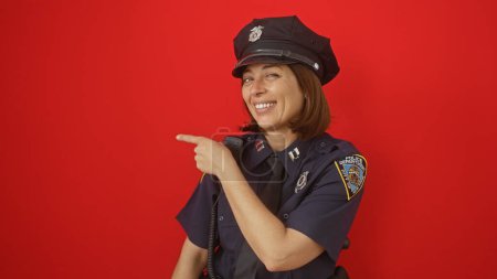 Smiling woman police officer in uniform pointing to the side against a red background