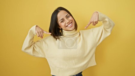 Foto de Mujer joven hispana sonriente en un suéter crema posando sobre un fondo amarillo, exudando confianza y alegría. - Imagen libre de derechos