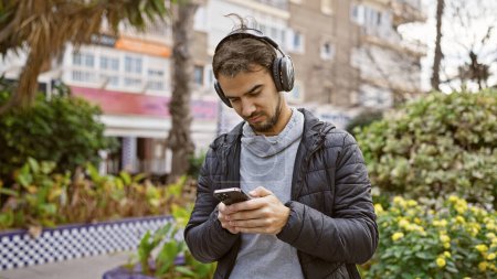 Foto de Un joven hispano con barba, con auriculares, mira su teléfono en un exuberante parque de la ciudad. - Imagen libre de derechos
