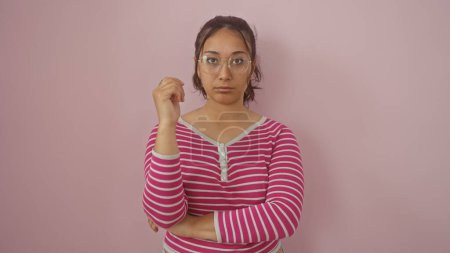 Foto de Mujer hispana joven y confiada con gafas y una camisa a rayas se levanta sobre un fondo rosado, exudando belleza y aplomo. - Imagen libre de derechos