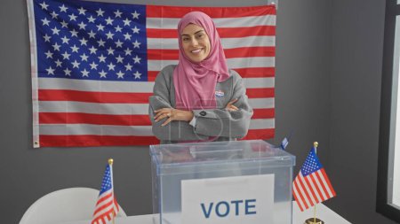 Una joven mujer hispana sonriente con un hiyab, de pie con confianza detrás de una urna en una sala de colegio electoral americana.